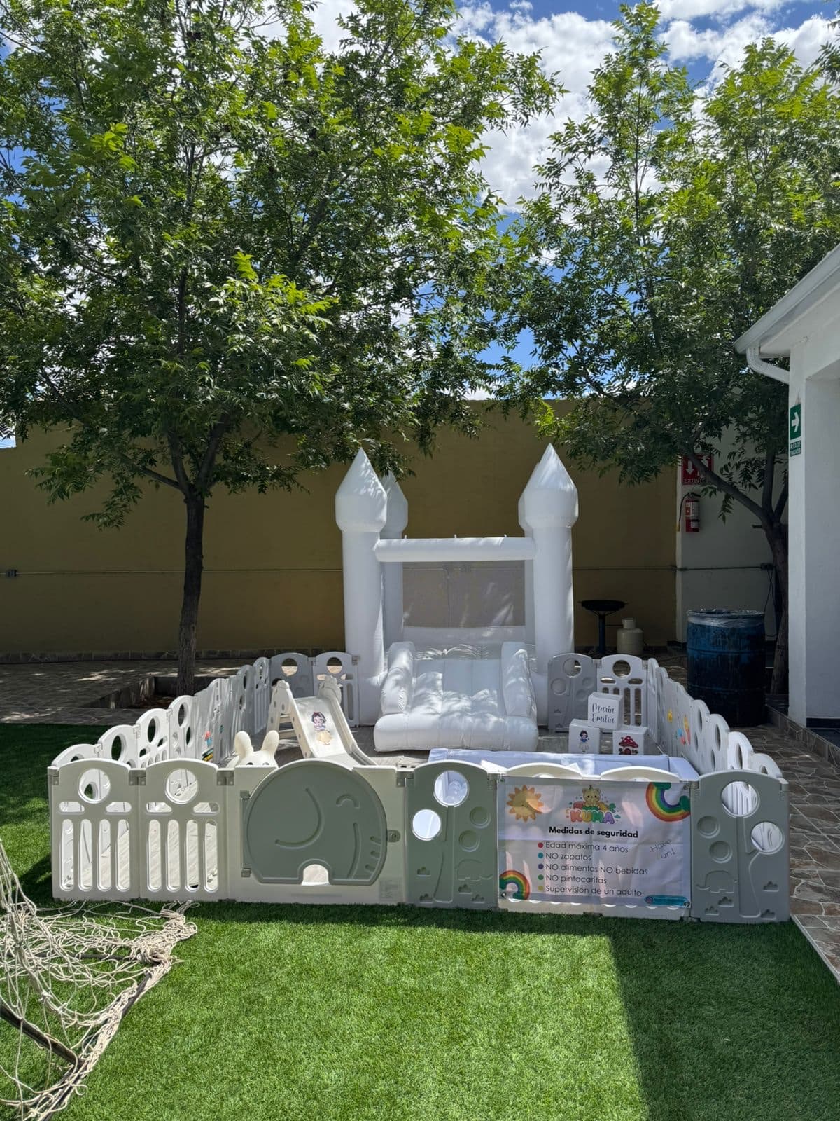 Setup completo del cumpleaños de María al aire libre en jardín con césped, árboles y pared blanca